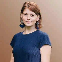 Woman with shoulder-length reddish-brown hair wearing a blue short-sleeved top and large earrings, standing against a plain beige background.