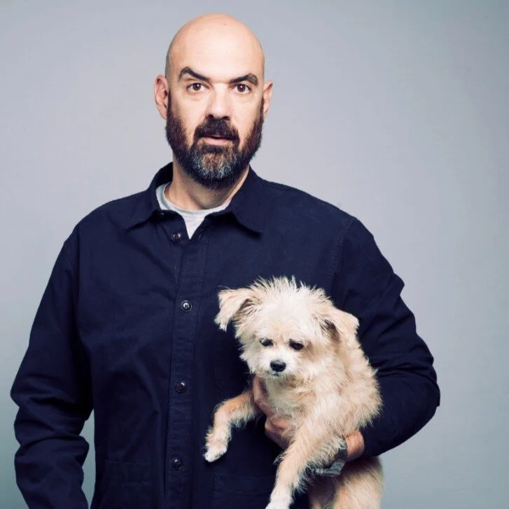 Man with a shaved head and beard wearing a dark button-up shirt, holding a small, fluffy light-colored dog against a plain grey background.