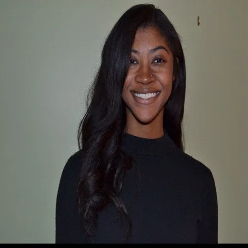 Woman with long, dark wavy hair wearing a black long-sleeved top, smiling and standing against a plain light-colored background.