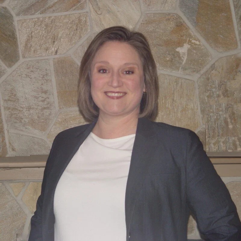Woman with shoulder-length light brown hair wearing a dark blazer over a light top, smiling while standing in front of a beige stone wall.