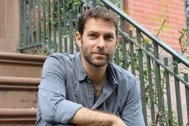 Man with short dark hair and beard wearing a blue button-up shirt, sitting on outdoor steps with a black railing and red brick wall in the background.