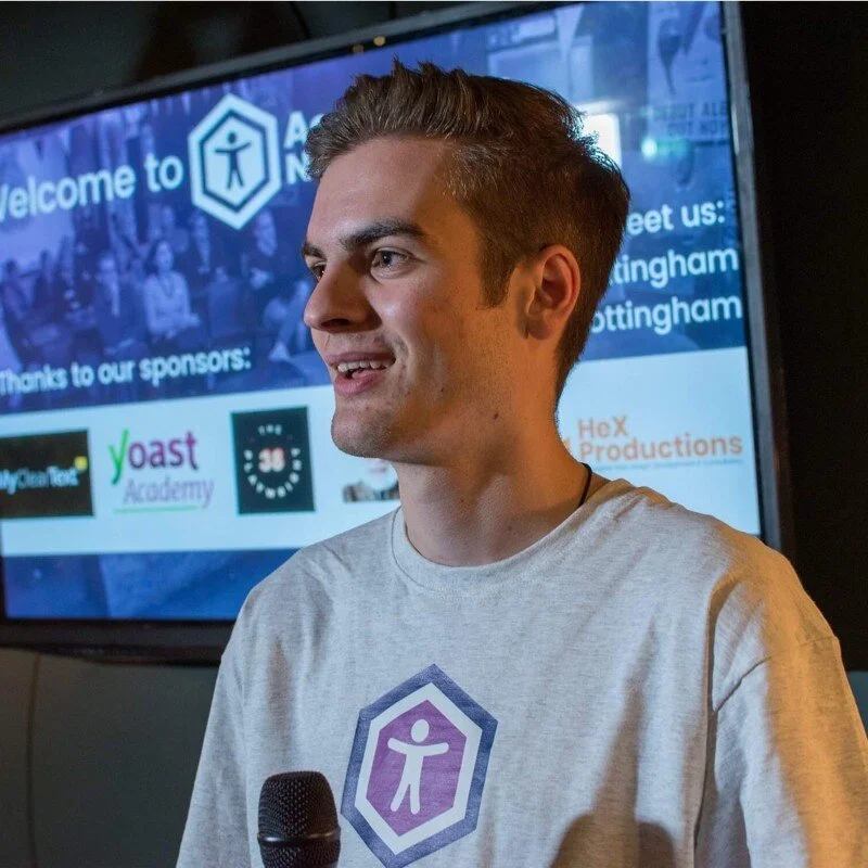 Man with short brown hair speaking into a microphone while smiling, wearing a light grey T-shirt with a purple and white logo, standing in front of a large screen displaying sponsor logos and event information.