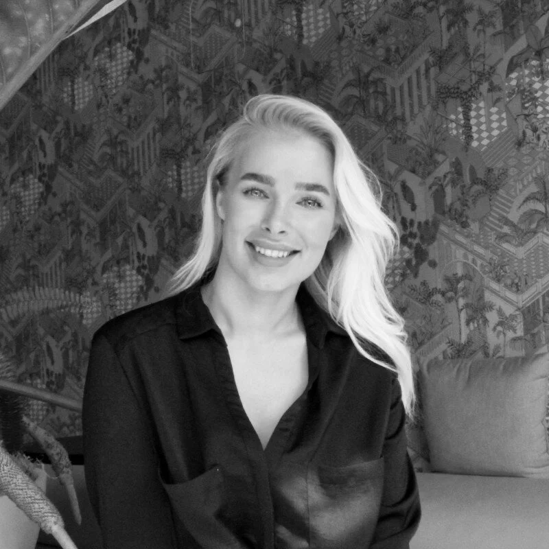Black and white photo of a woman with long, light-colored hair wearing a dark button-up shirt, smiling while seated in front of a patterned background.