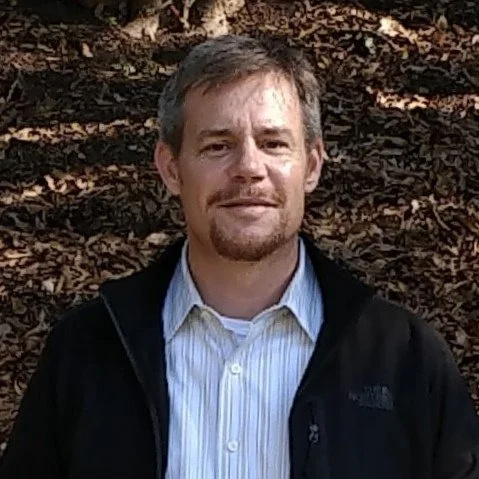 Man with short grayish hair wearing a black jacket over a light blue button-up shirt, standing outdoors in front of a background of fallen leaves.