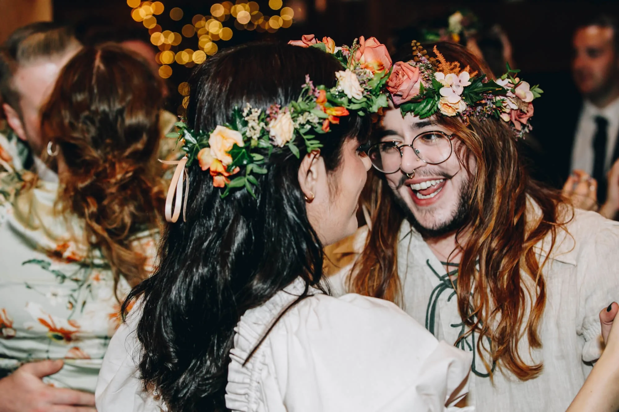 Two people laughing and celebrating at a wedding, wearing flower crowns with pink and peach roses and greenery, in a festive indoor setting with warm lighting and blurred background of other people.