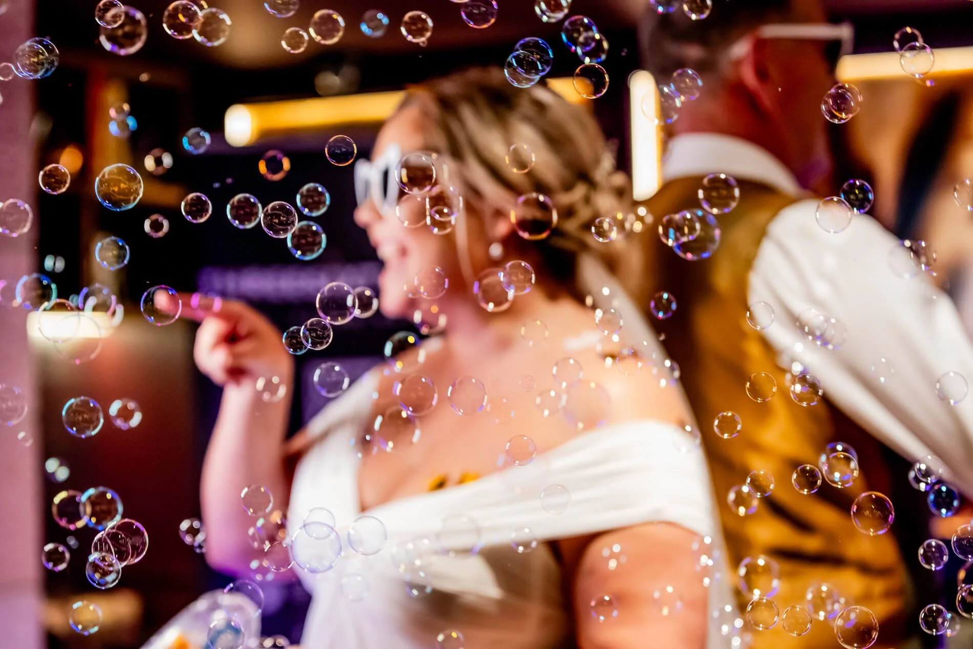 A woman with short hair wearing sunglasses and a white off-shoulder dress smiling and pointing. A man in a yellow jacket and sunglasses is beside her. There are soap bubbles floating all around them in an indoor setting with warm lighting.