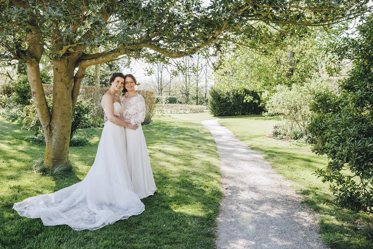 Two women in wedding dresses standing under a tree on a sunny day in a lush green garden