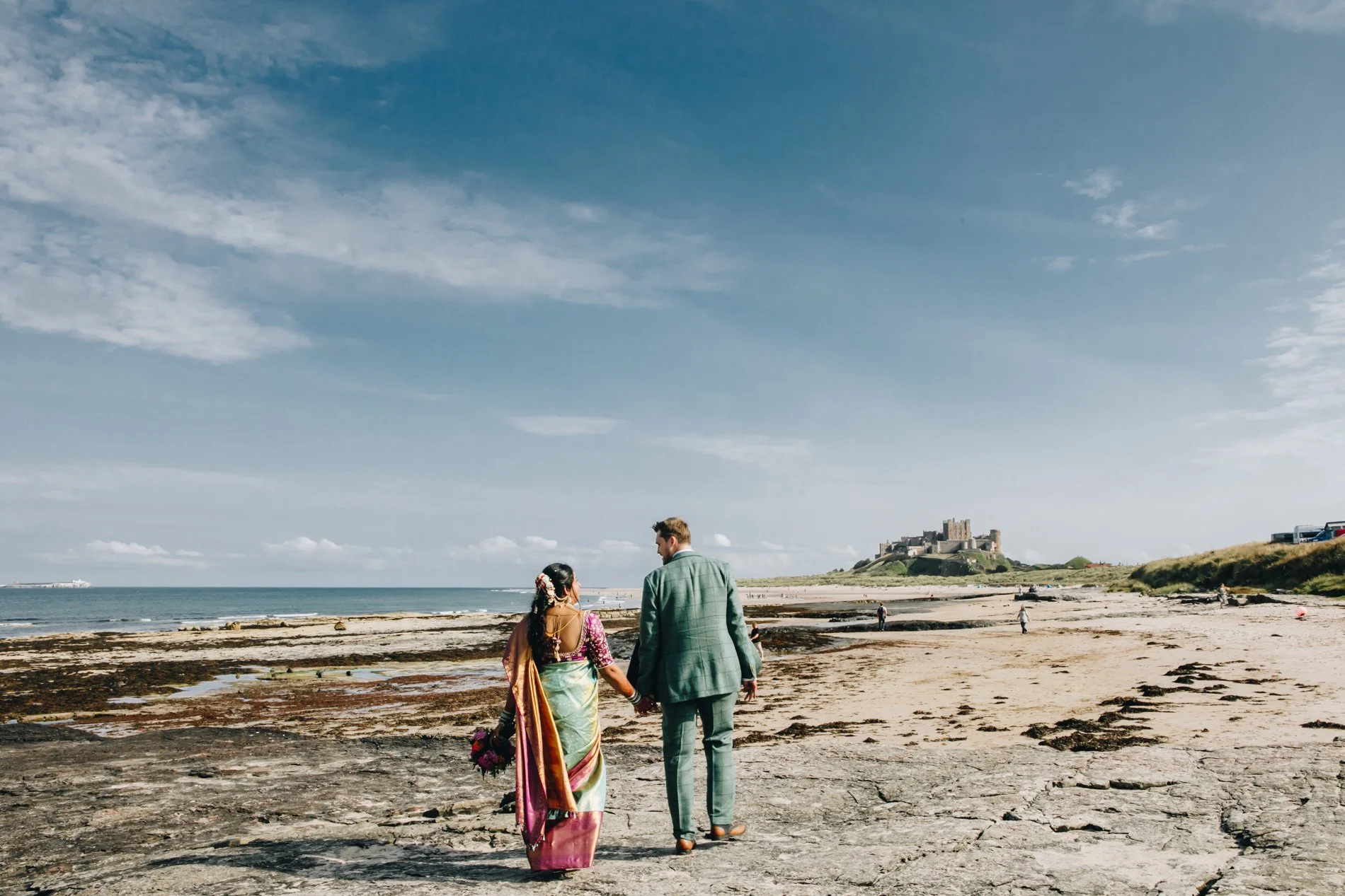 Newlyweds walking hand-in-hand along a rocky shoreline towards Bamburgh Castle under a blue sky, capturing a natural and quiet moment