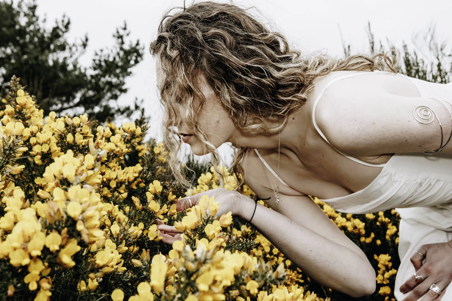 A woman with curly hair and jewellery, leaning over yellow flowers outdoors, closely looking at gorse flowers