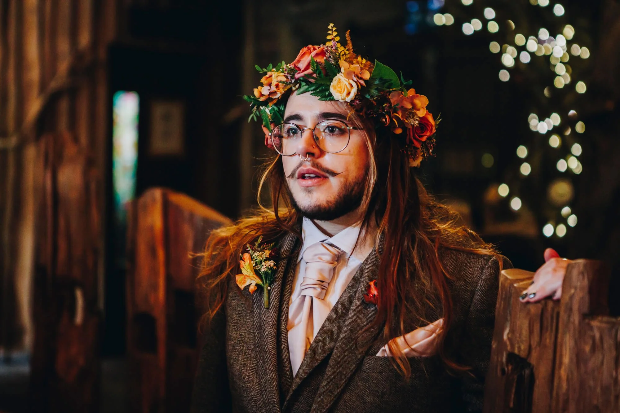 A groom with long hair, glasses, and a septum piercing, wearing a floral crown and suit, looking at the room where his wedding breakfast will take place at Alnwick Treehouse. He is emotional. 