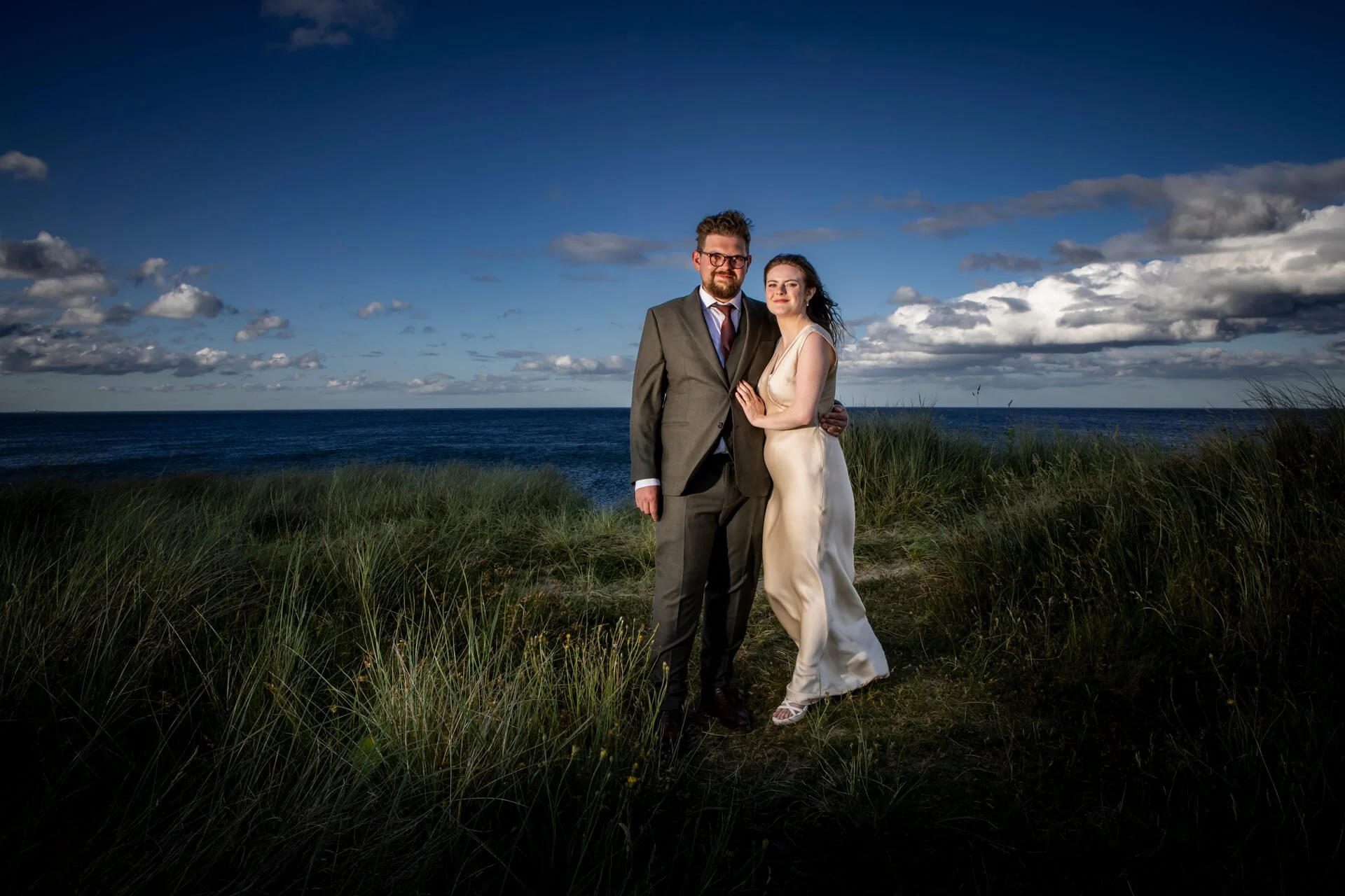 A man and woman in formal wedding attire standing on a grassy area near the ocean, with a cloudy sky above.