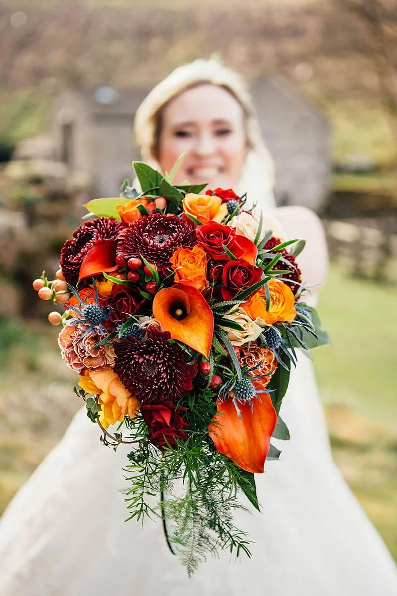 A bride in her wedding dress holding a colourful red, orange and yellow bouquet of flowers outdoors, with a blurred background of trees. The Priests House North Yorkshire