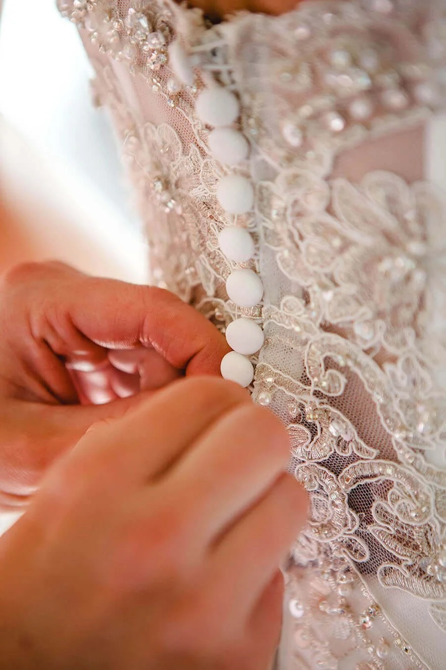 Close-up of a person buttoning the back of an elegant, pale pink or blush wedding dress with intricate lace and beadwork.