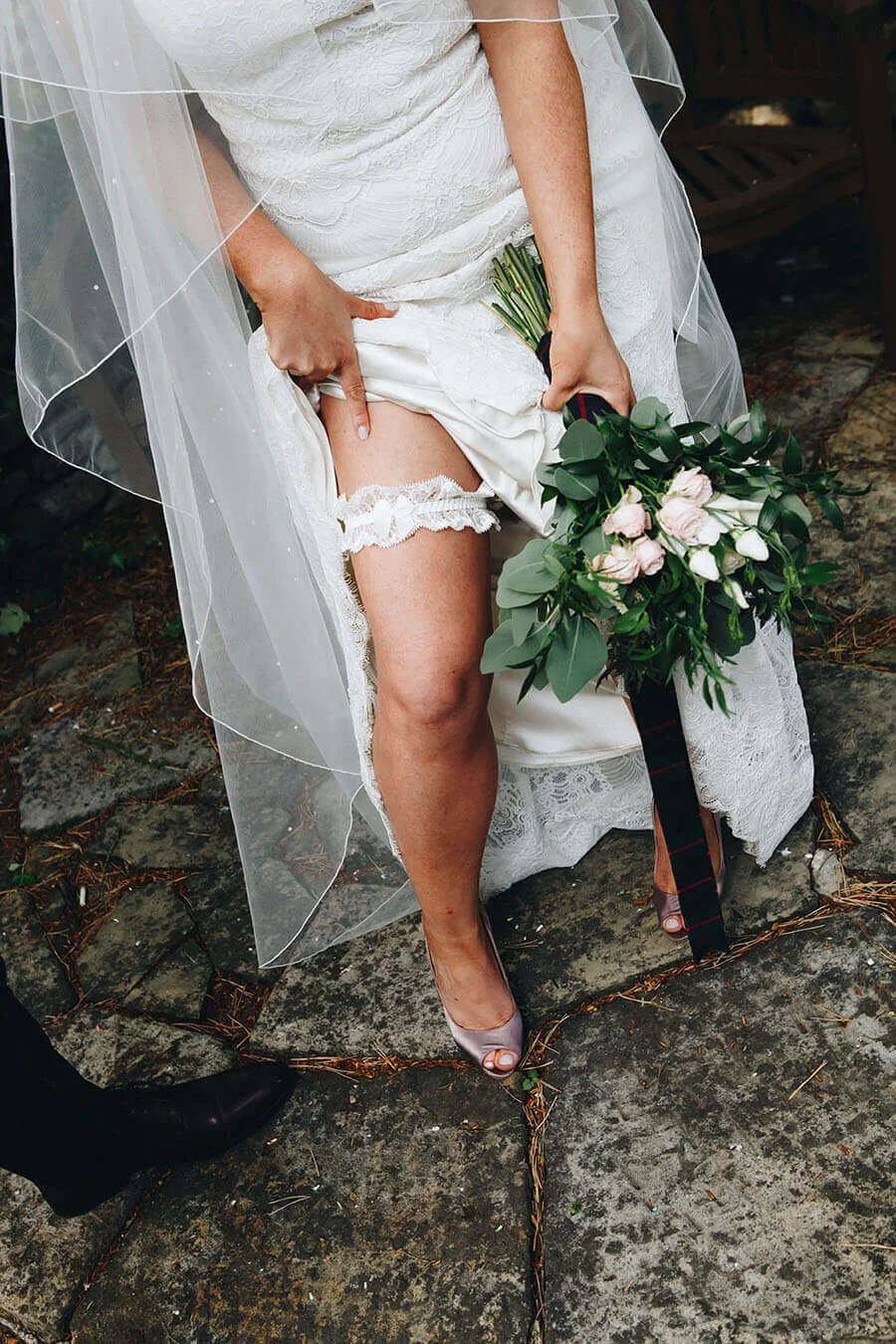 Bride lifting her wedding dress to show her lace garter while holding a bouquet of pink and white flowers, standing on a stone patio outdoors.