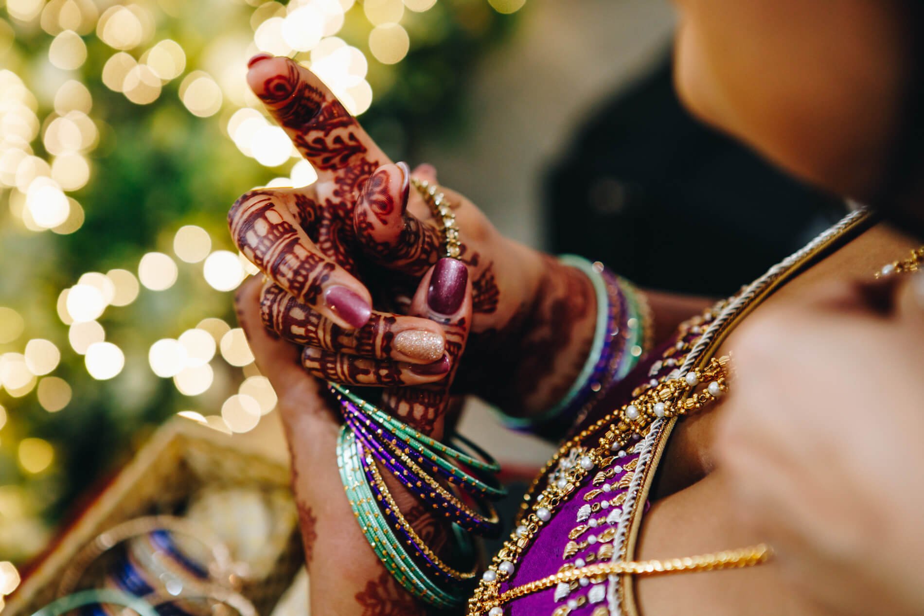 Close-up of a woman with intricate henna designs on her hand, wearing colorful bangles, jewelry, and a deep purple traditional dress, with warm bokeh lights in the background at Bamburgh Castle