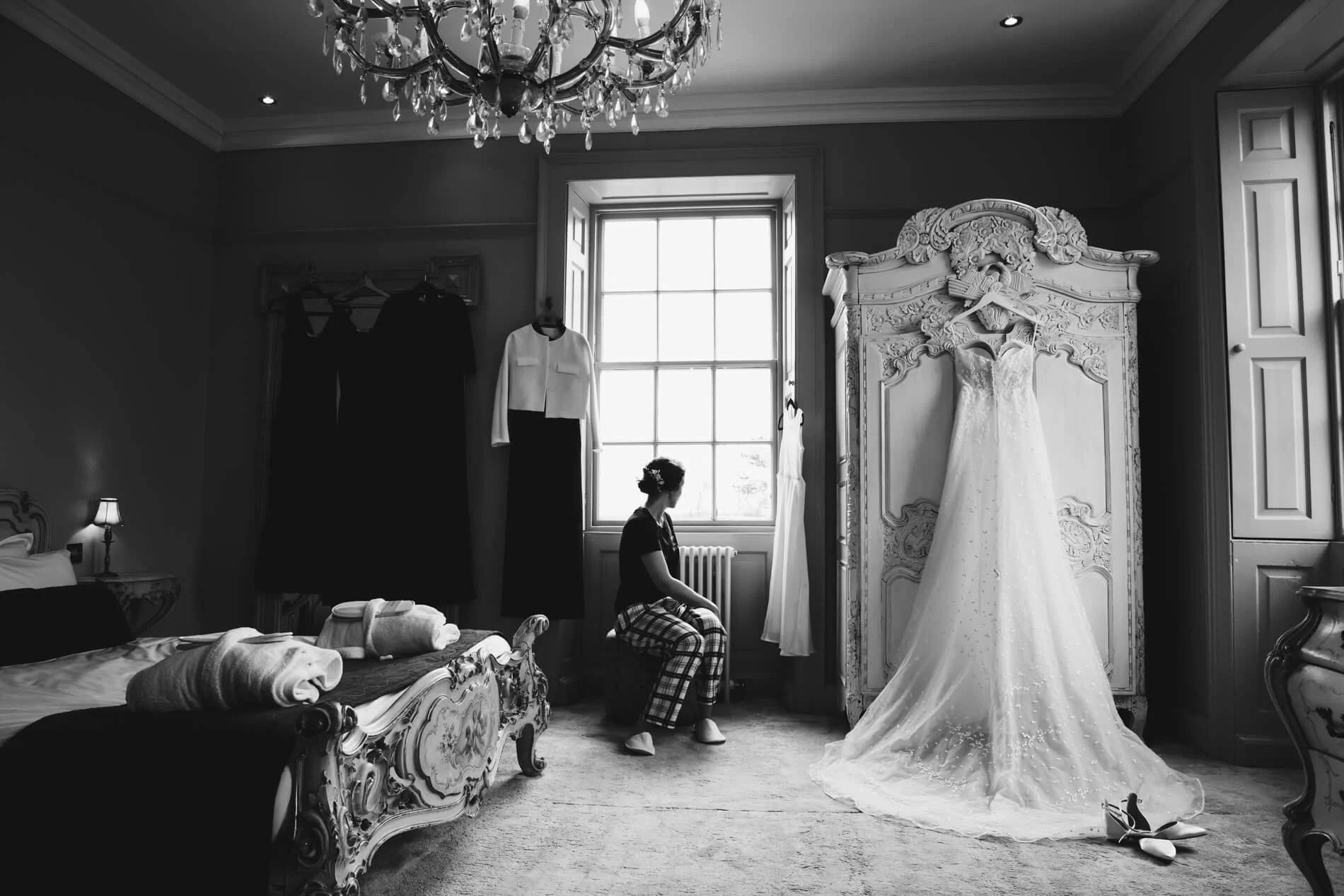 A woman sits on a window seat in an ornate, vintage bedroom with a large, decorative wardrobe and clothing hanging on a rack. A wedding dress hangs on the wardrobe, and a pair of shoes is on the floor nearby. The room is illuminated by natural light 