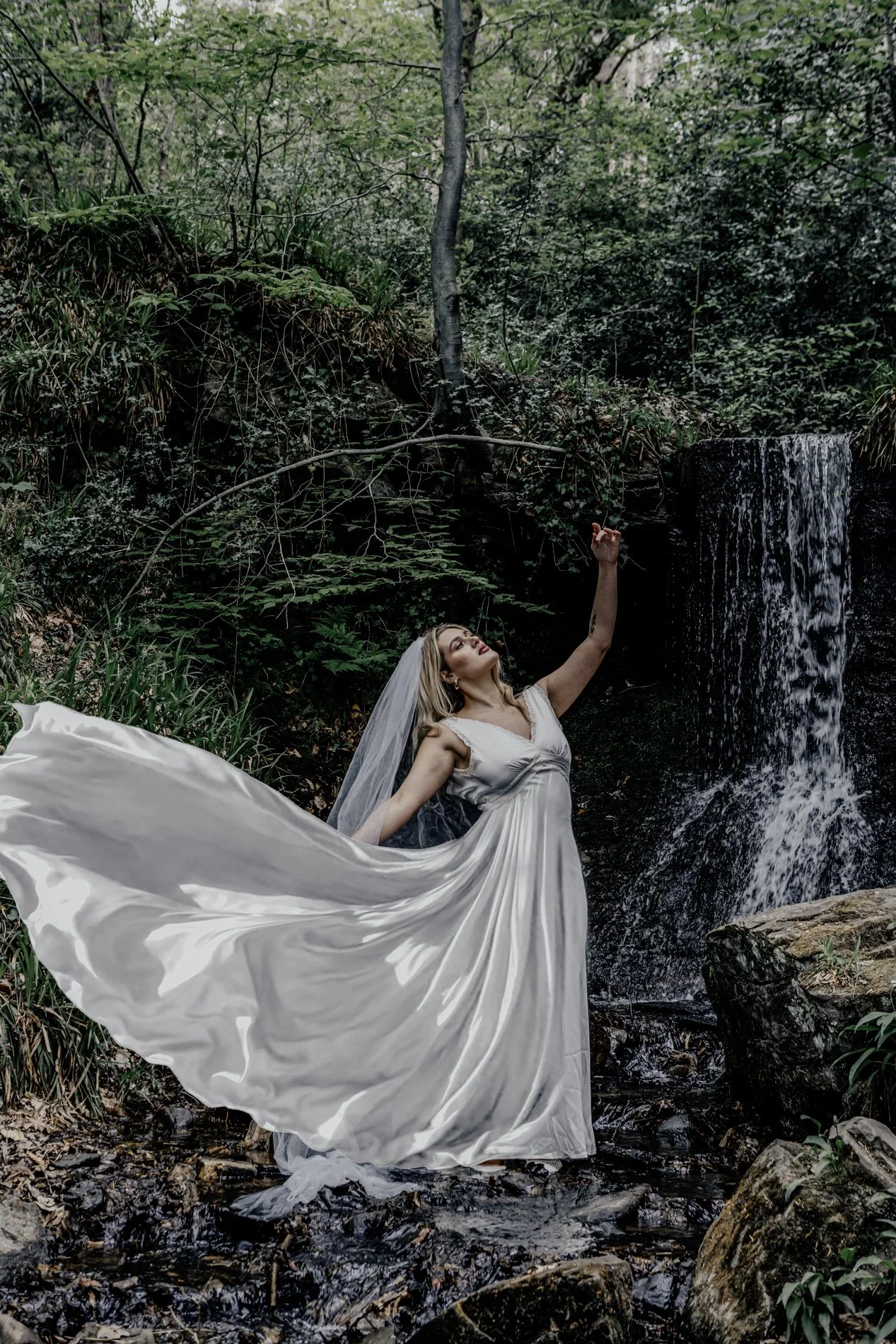 A woman in a white flowing dress and veil standing on a rocky stream near a waterfall, surrounded by lush green foliage, striking a graceful pose.