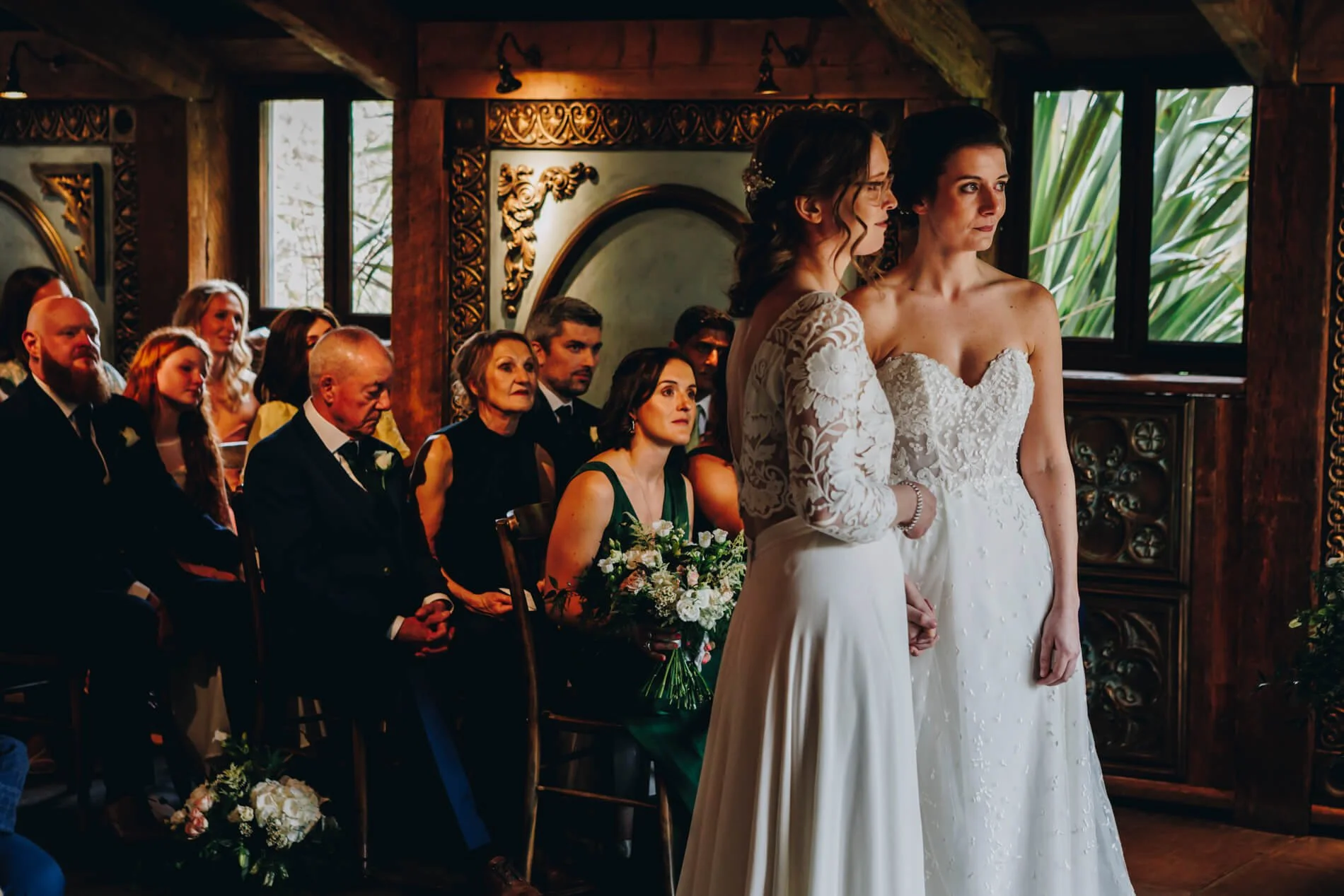 Two brides in wedding dresses holding hands during a ceremony, surrounded by seated guests in a decorated indoor venue with wood walls and windows. Newton Hall