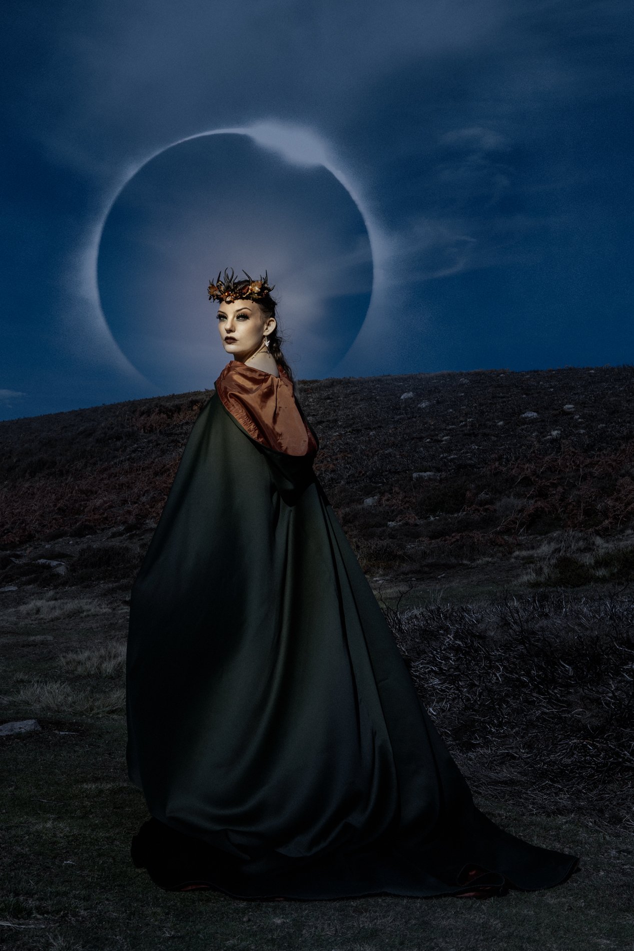 A woman wearing a dark green cloak with a red satin shoulder detail stands outdoors under a moon with a solar eclipse visible in the sky.