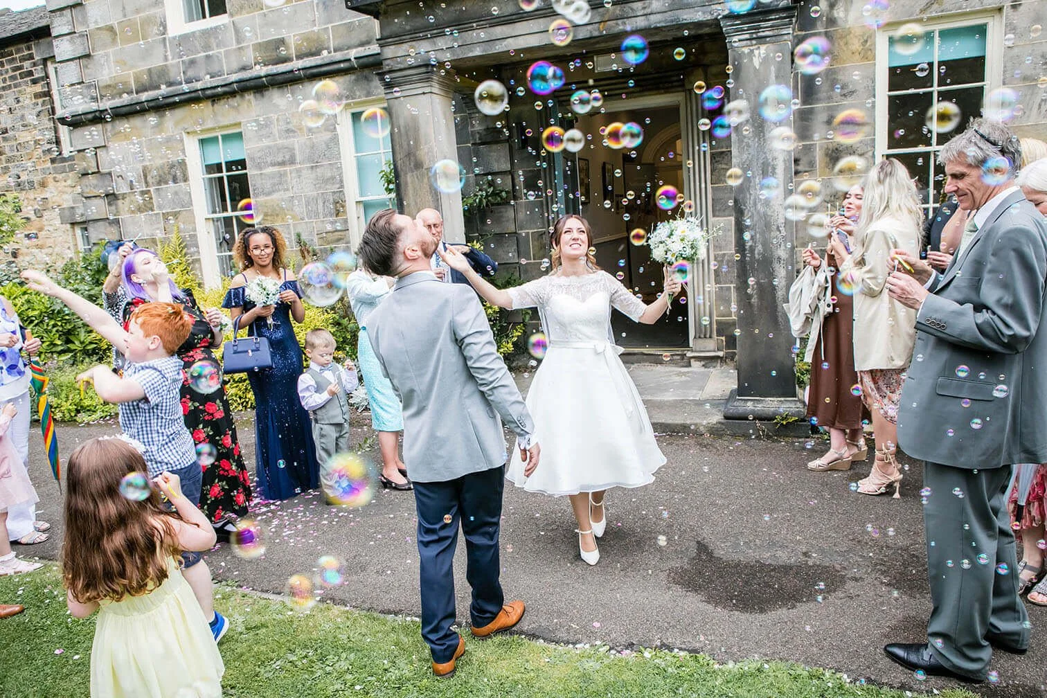 Bride and groom celebrating with guests outside a building, surrounded by bubbles and flower petals, during a wedding reception. Hexham Registry Office