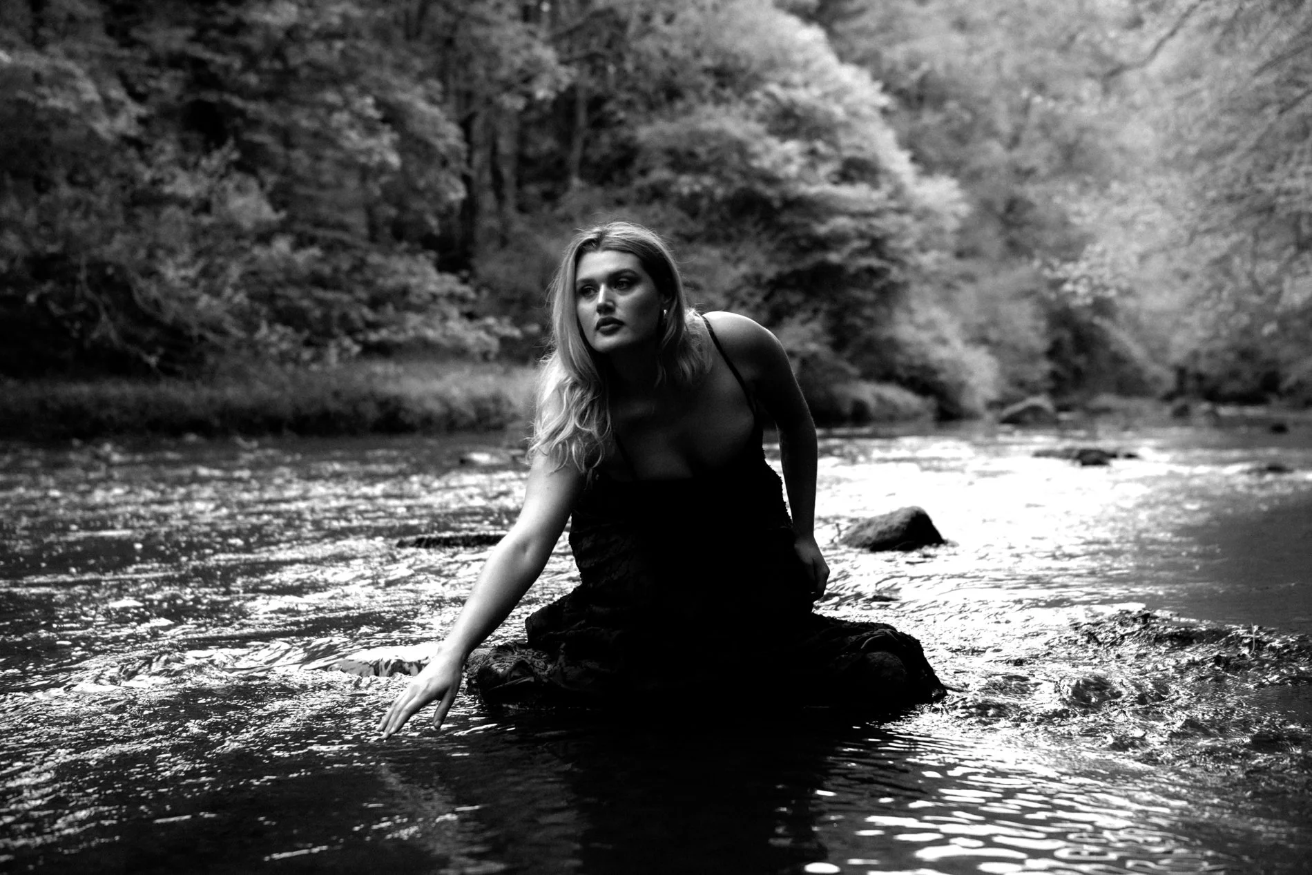 A woman with wavy hair in a black dress kneels in a river, surrounded by trees, in black and white.