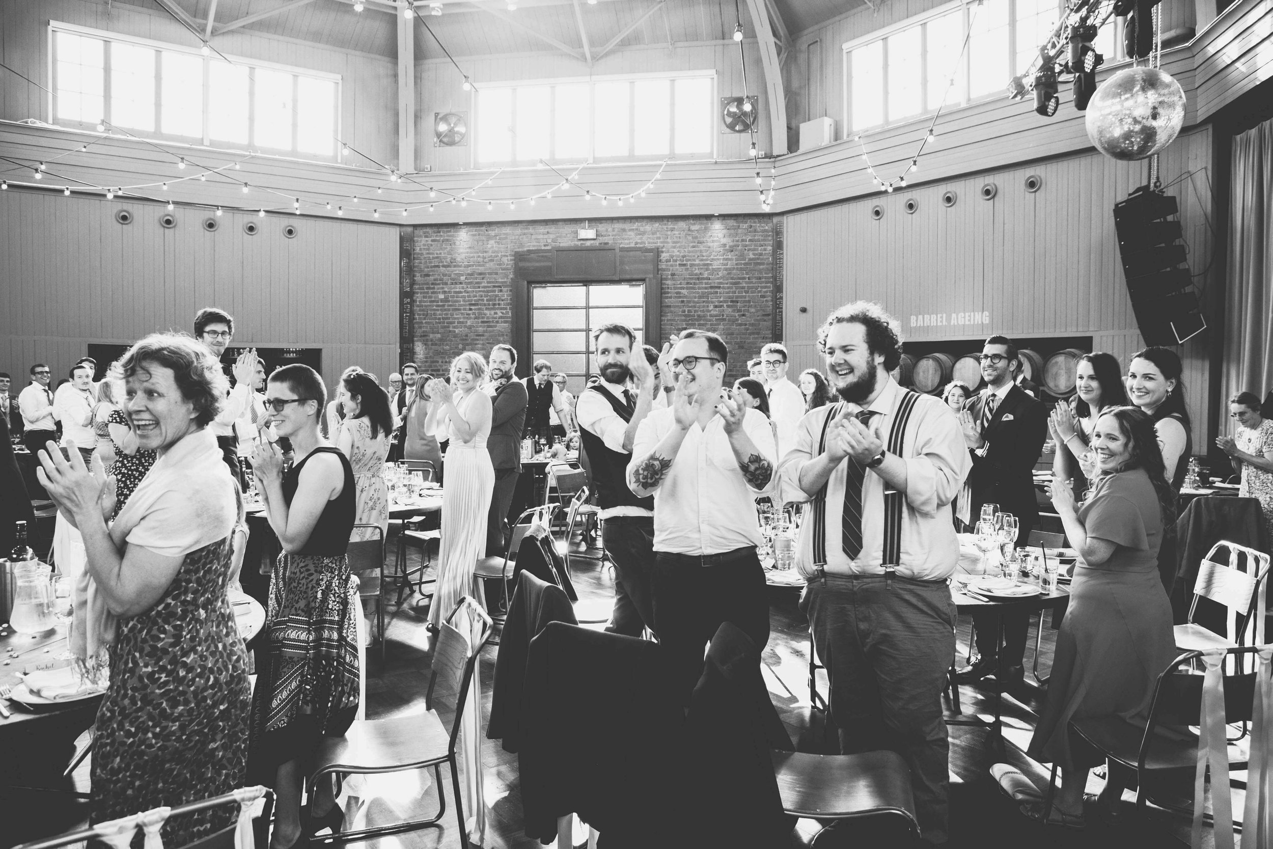 People standing and clapping in the decorated event hall at Wylam Brewery, some smiling and laughing during a wedding.