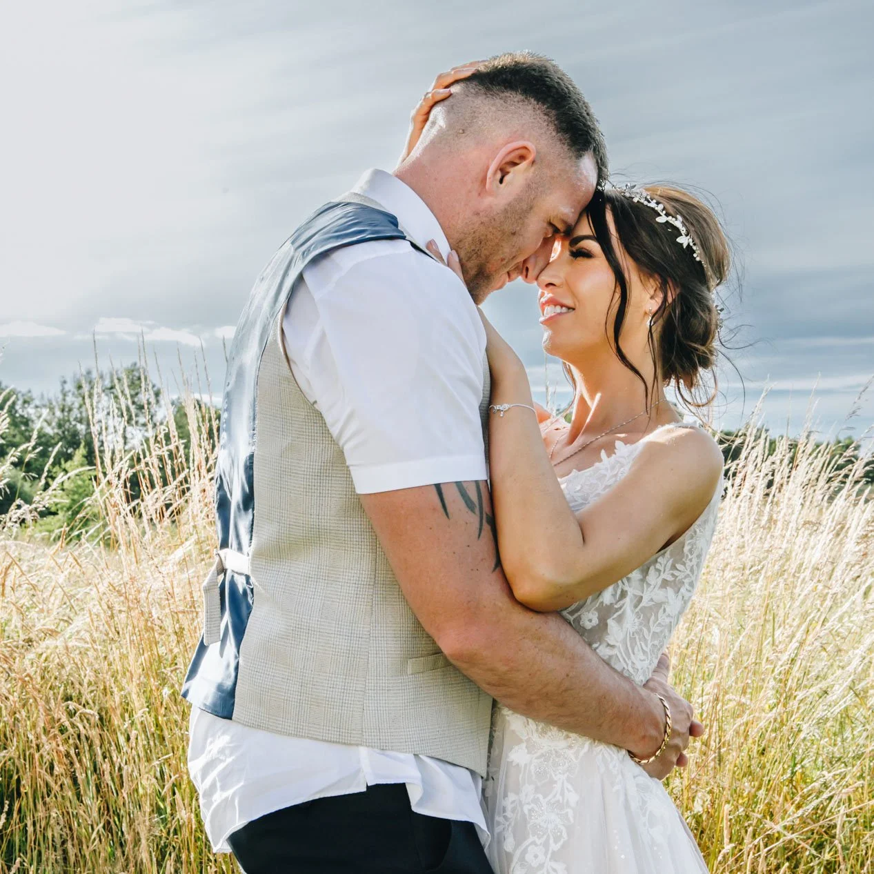 A newlywed couple embraces affectionately outdoors in a field of tall grass, with a cloudy sky in the background.