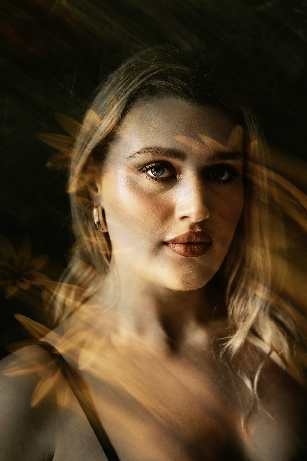 A woman with blonde hair and makeup posing against a dark background with blurred orange flower reflections.