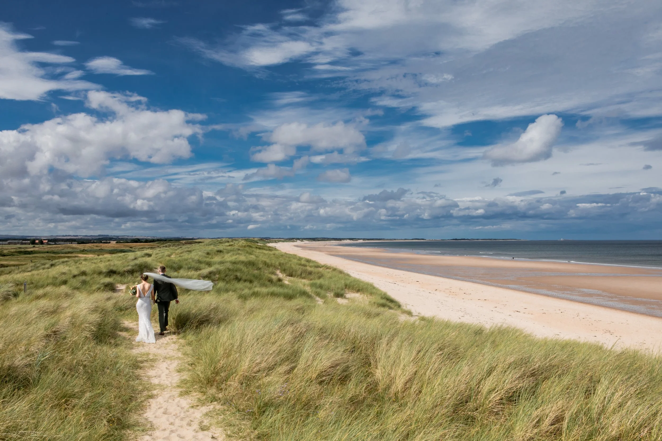 A bride and groom walking through the tall grass of the sand dunes overlooking a vast, empty beach on the Northumberland coast