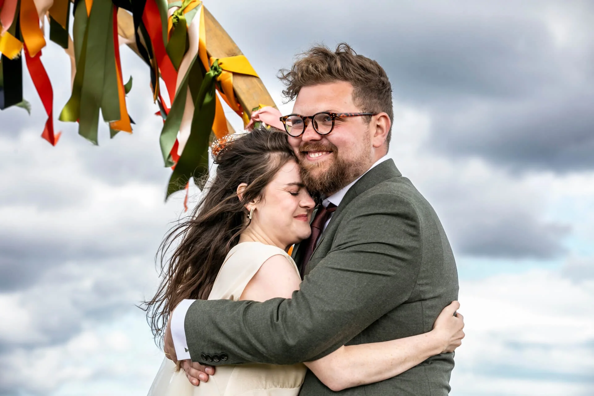 A couple hugging outdoors during a cloudy day, with colourful ribbons hanging above them. The Barn on the Bay, outdoor wedding