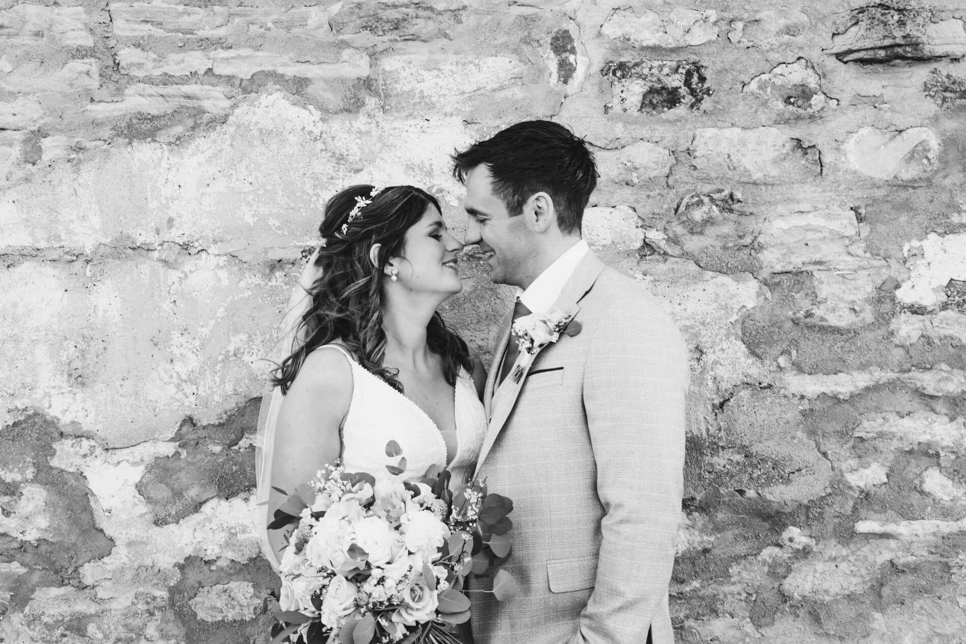 A newlywed couple in wedding attire standing close together with foreheads touching, smiling, and holding a bouquet of flowers, against a textured brick wall background.