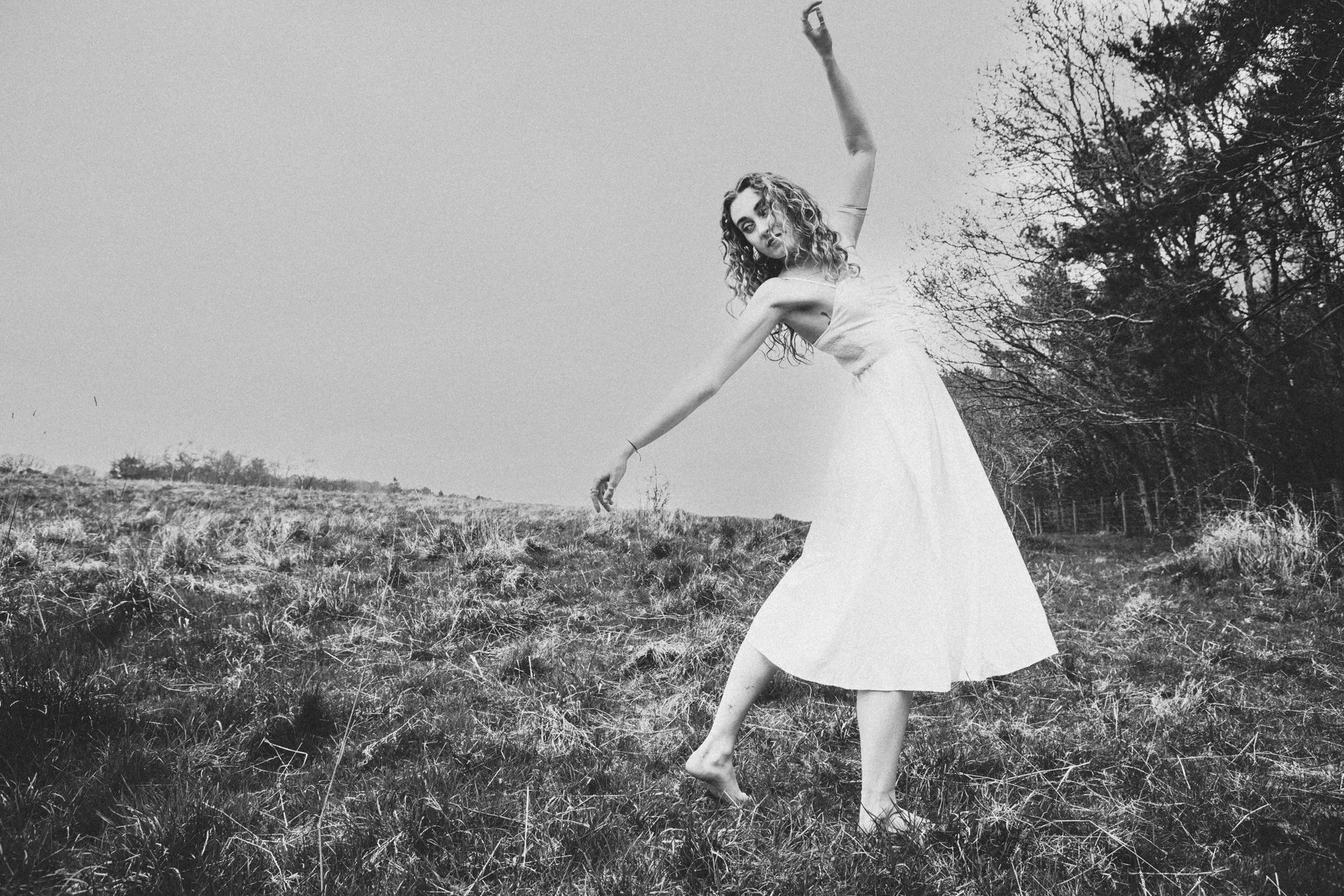 A woman wearing a sleeveless dress dancing barefoot outdoors on grassy terrain with a backdrop of trees and an open sky, captured in black and white.