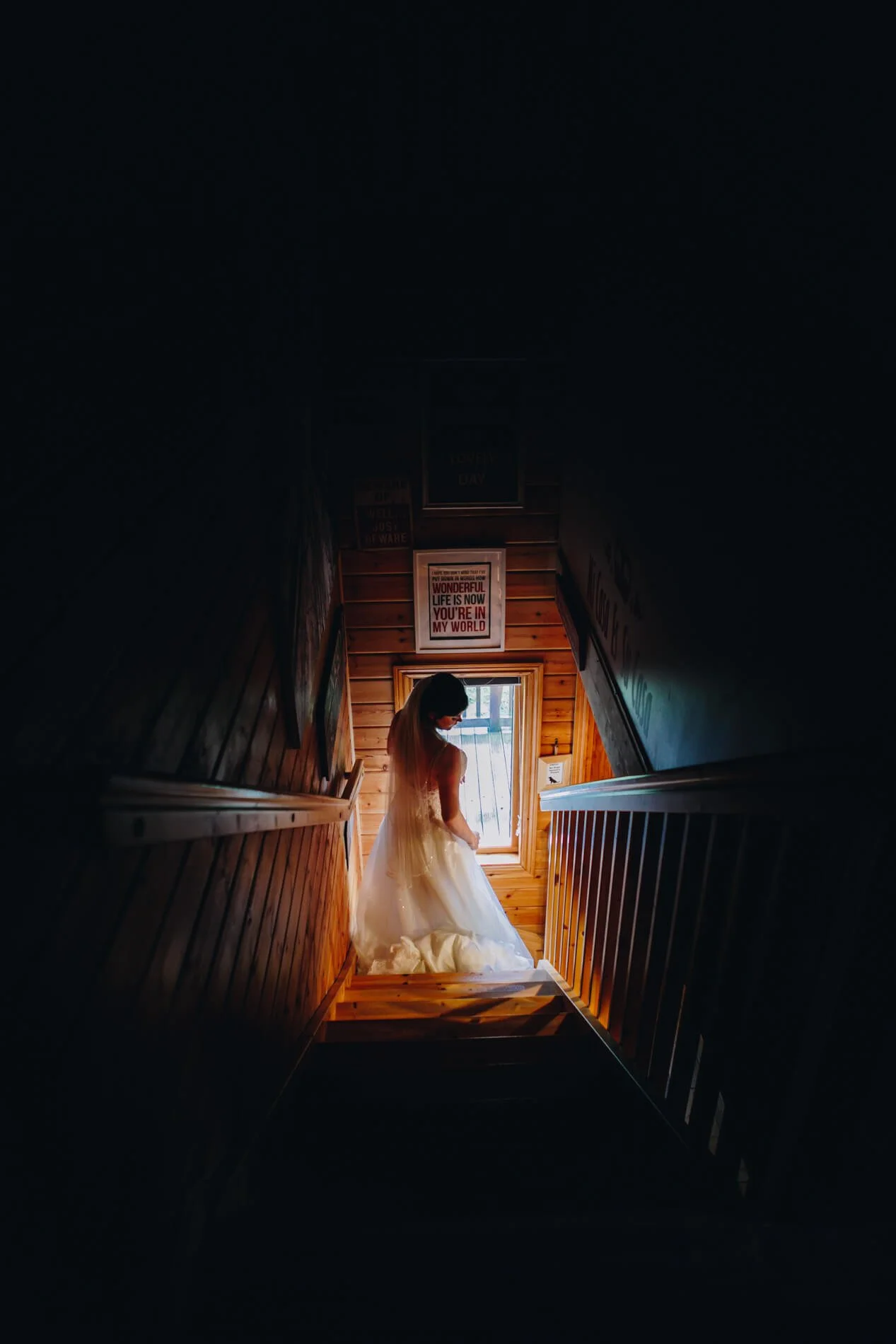A bride in a wedding dress on a staircase near a window in a wooden interior.