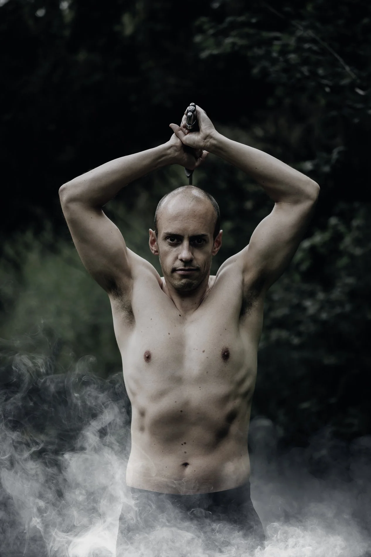 A shirtless man wielding a sword over his head in a smoke-filled wood