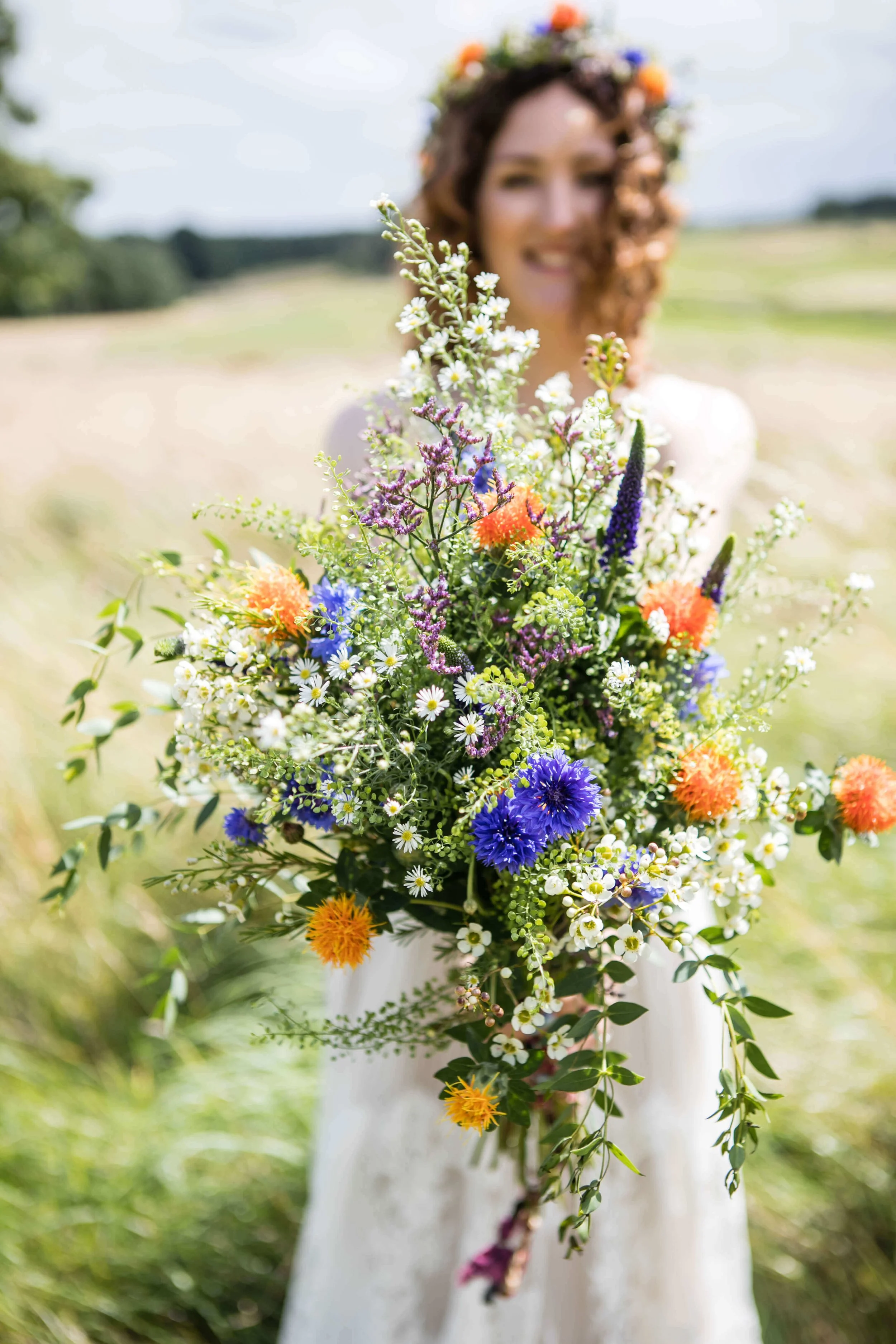 A woman with curly hair wearing a floral crown holds a large bouquet of colourful wildflowers outdoors in the rough at Matfen Hall on the golf course