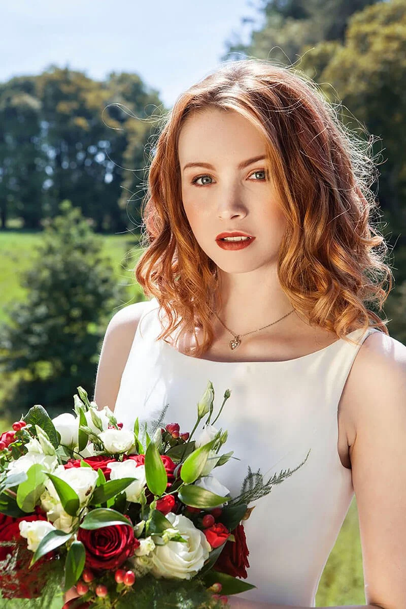 A young woman with curly red hair in a sleeveless white dress holds a bouquet of red and white flowers outdoors. She has red lipstick and a heart-shaped pendant necklace, with a background of green trees and blue sky.