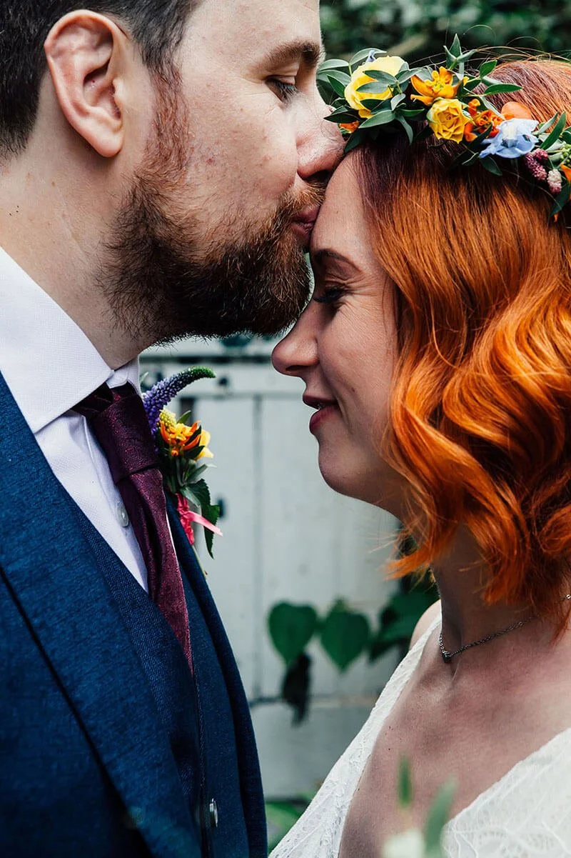 A couple on their wedding day with foreheads touching, eyes closed, with the man kissing the woman's forehead. The man has a beard and is wearing a blue suit with a white shirt and dark tie. The woman has red hair with waves and is wearing a floral c
