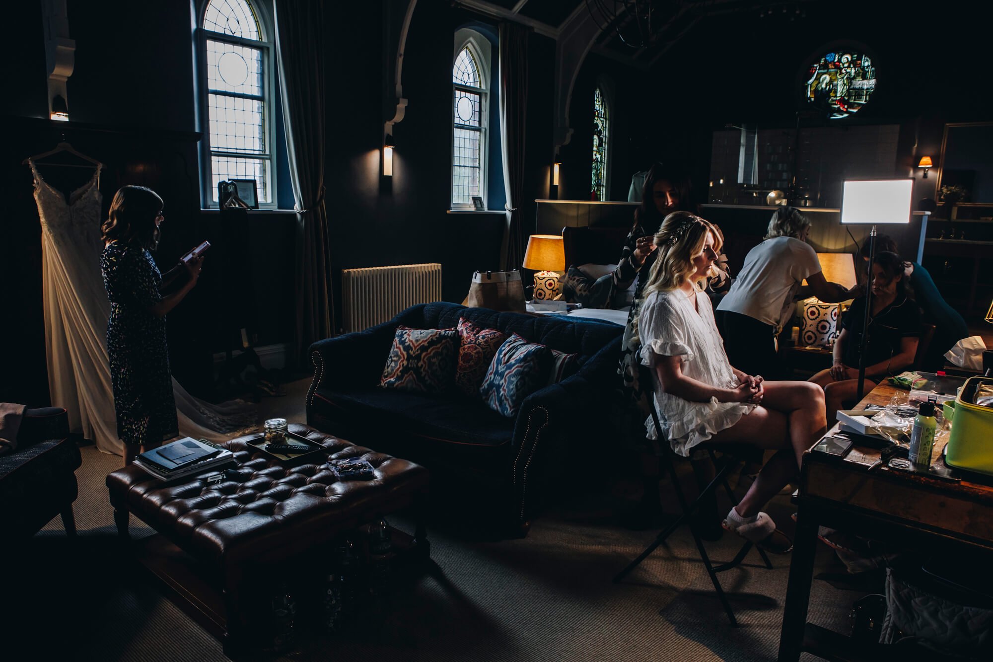 Interior of a dimly lit room with large arched windows, where several women are preparing, including one sitting on a chair with a white dress, while others work around her with makeup and hair tools, and a woman standing on the left using her phone.