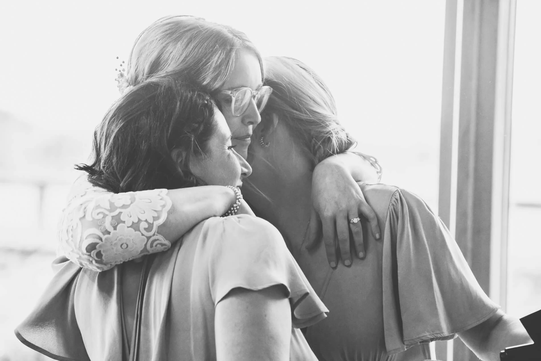 Three women embrace each other in a heartfelt hug by a window, with soft light illuminating their expressions of comfort and affection.
