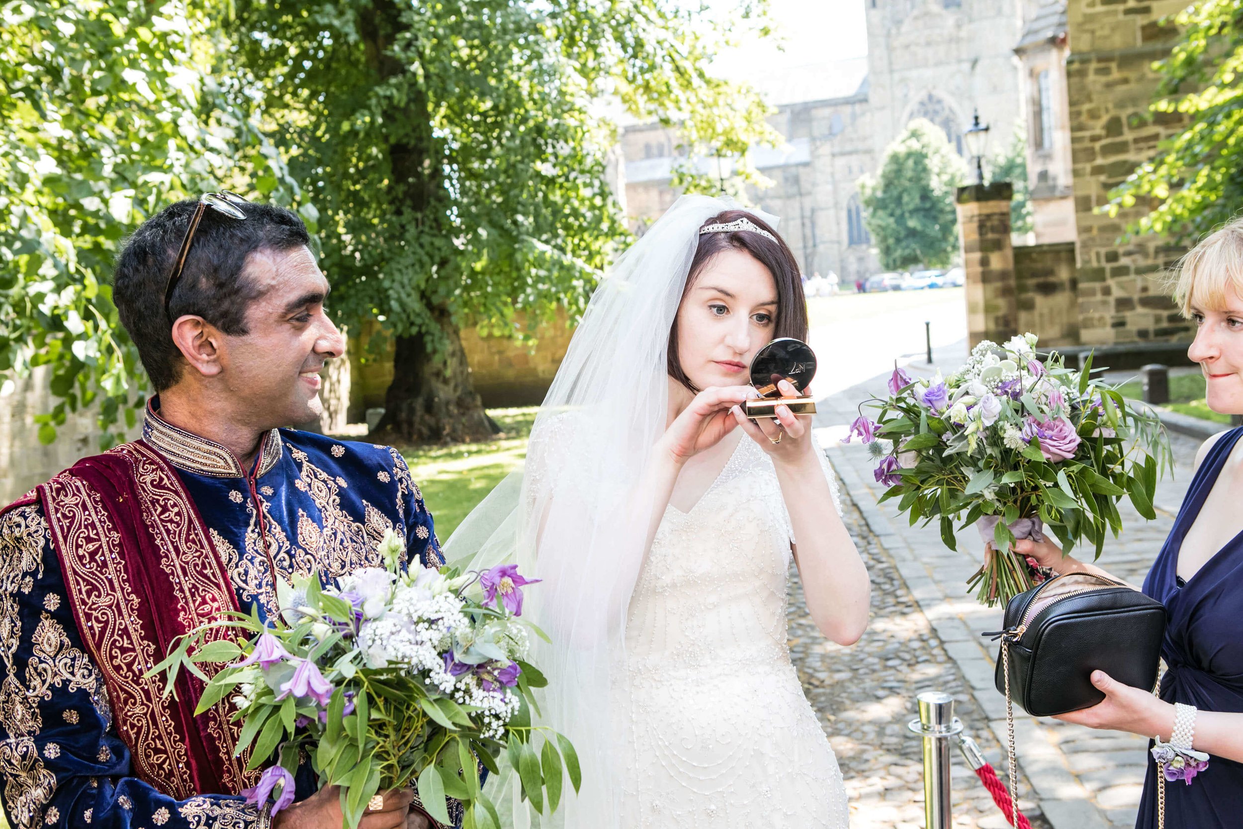 A bride getting ready for her wedding outdoors, holding a makeup compact mirror, with a man in traditional attire holding a bouquet of flowers, and a woman handing her a bouquet of flowers with historic Durham Castle in the background.