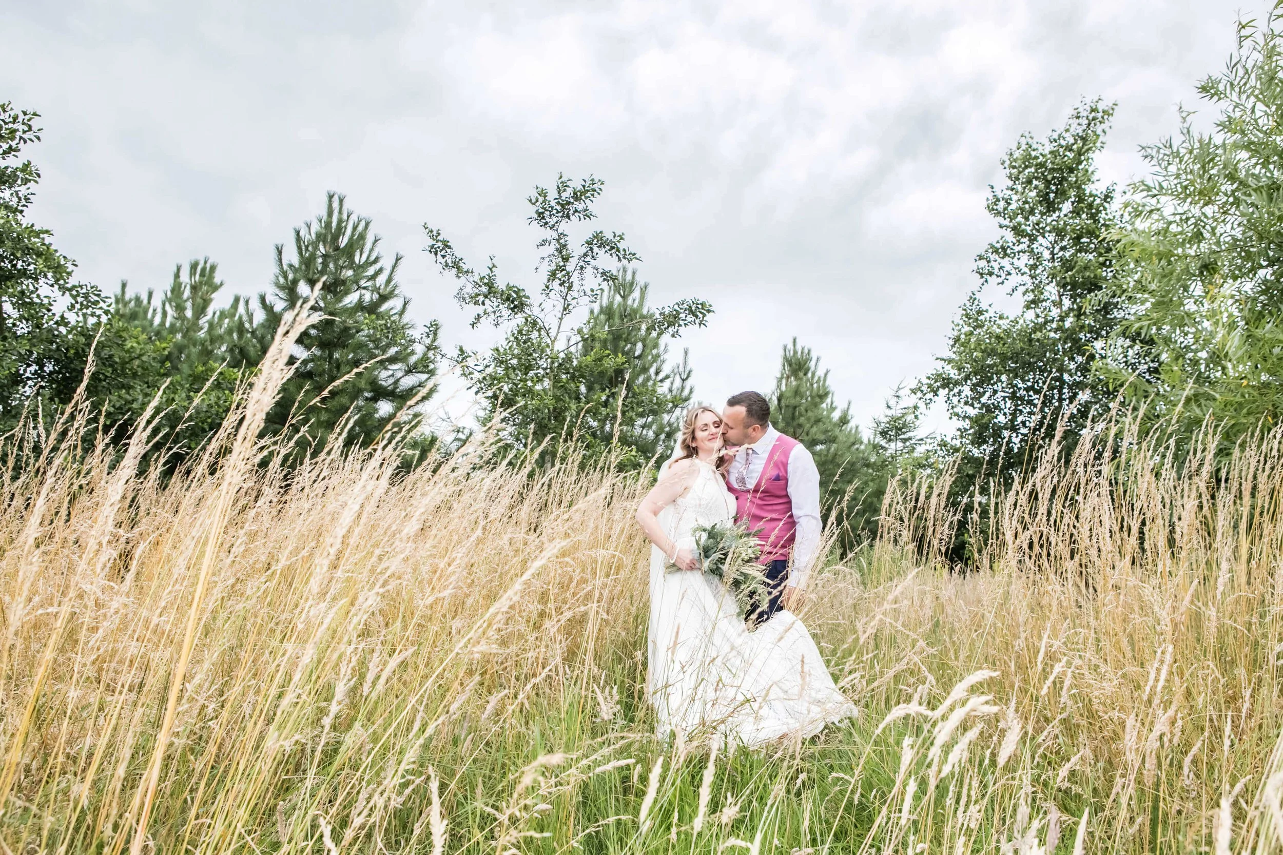 A couple in wedding attire standing in a grassy field, with the groom kissing the bride on the cheek. The bride is holding a bouquet, and tall grasses and trees are visible in the background under a cloudy sky. The wedding was at Vallum Farm
