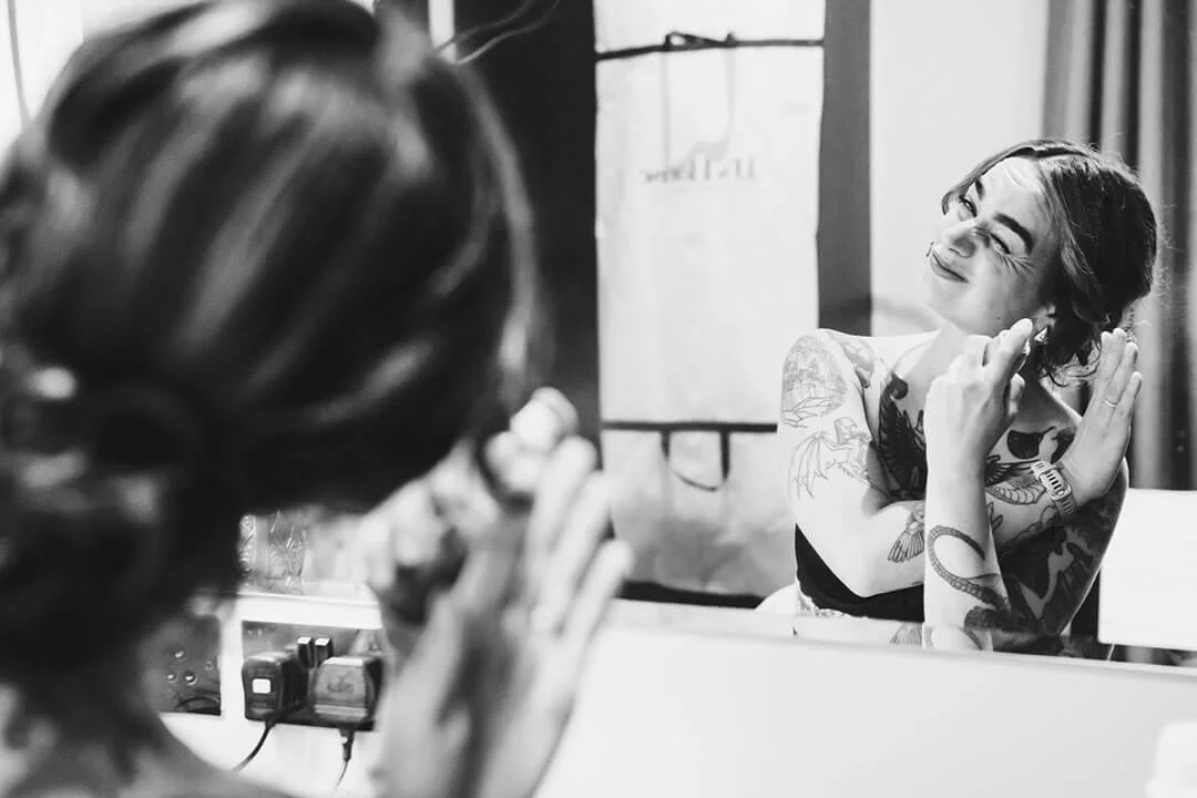 A woman with tattoos on her arms and shoulder, smiling and looking at herself in a mirror, adjusting her hair in a brightly lit room.