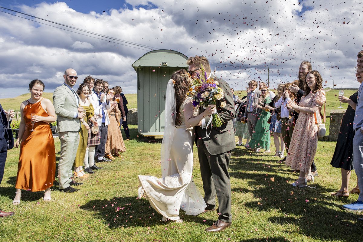 A wedding couple kisses while walking on grass, surrounded by guests throwing confetti on a cloudy day.