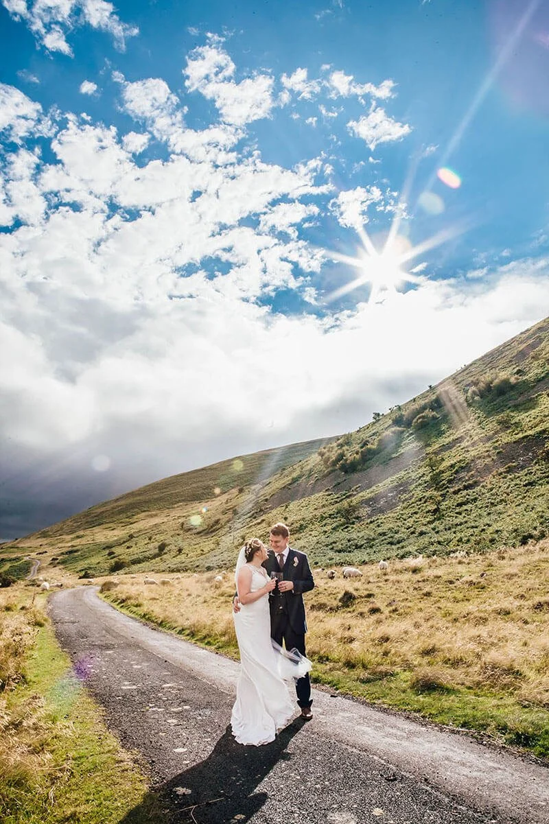 A bride and groom standing together on a dirt road in a hilly outdoor landscape under a bright sky with scattered clouds and the sun shining. College Valley, Cuddystone Hall wedding photography