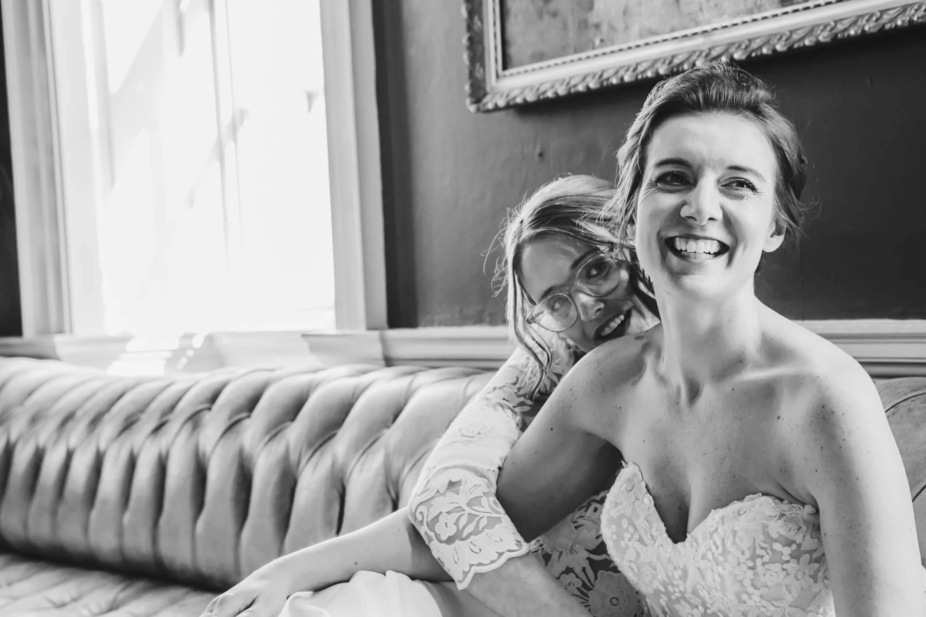 Two brides on their wedding day. One in a strapless wedding dress, smiling and looking away, sitting on a vintage tufted sofa next to another woman with glasses, smiling and leaning in close, indoors with natural light