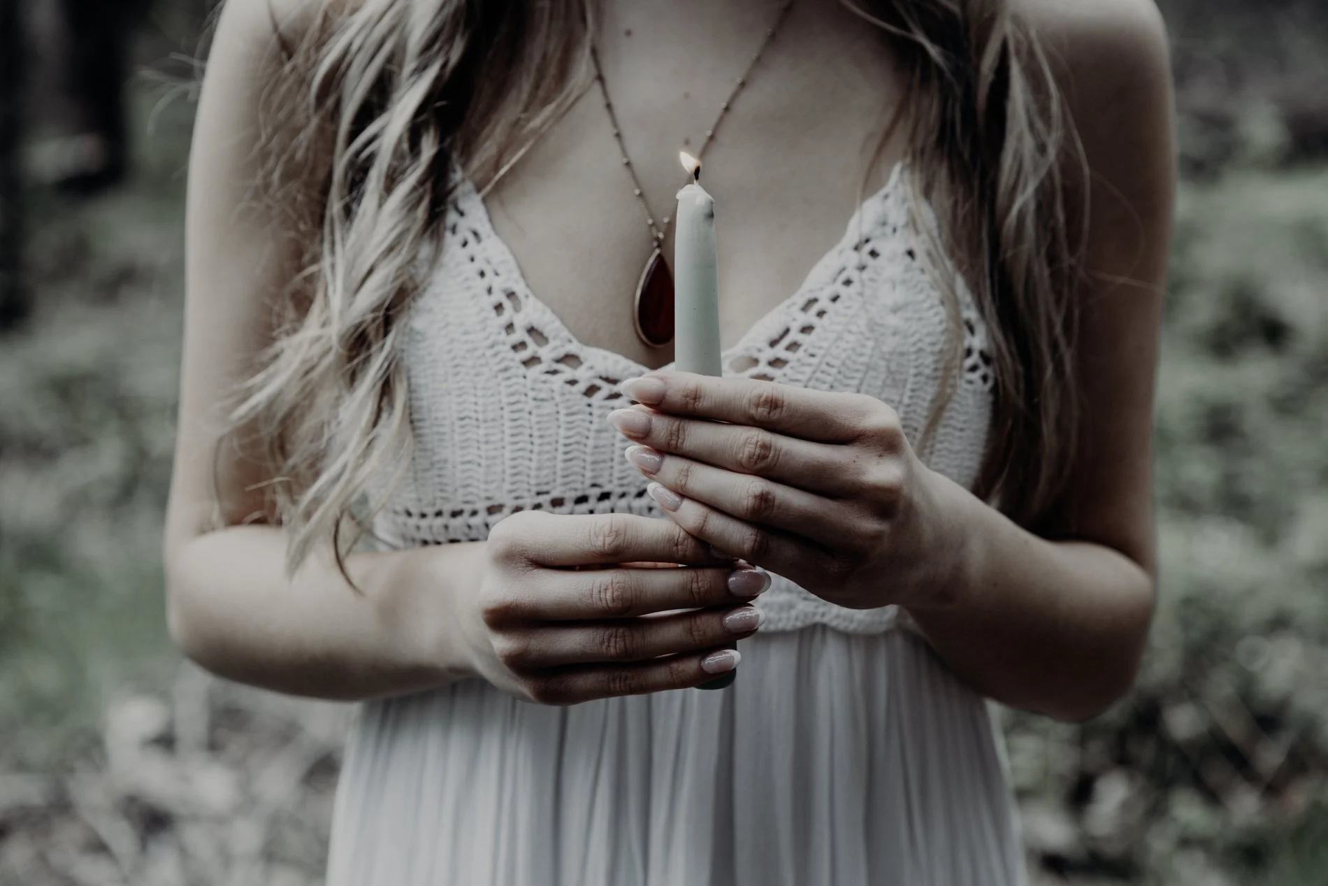 A woman wearing a white crochet dress holds a lit candle with both hands in an outdoor setting.