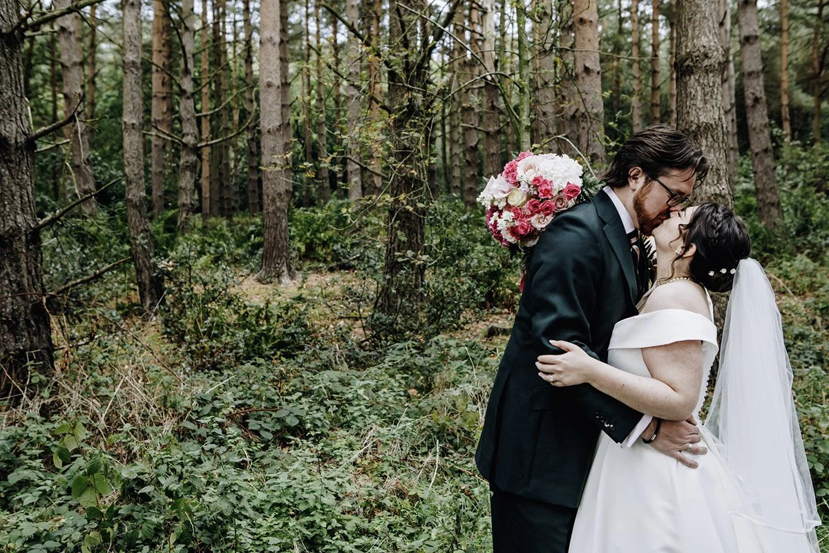 A bride and groom kissing in a forest, with the groom holding a bouquet of pink and white flowers, the bride wearing a veil, and both dressed in wedding attire.