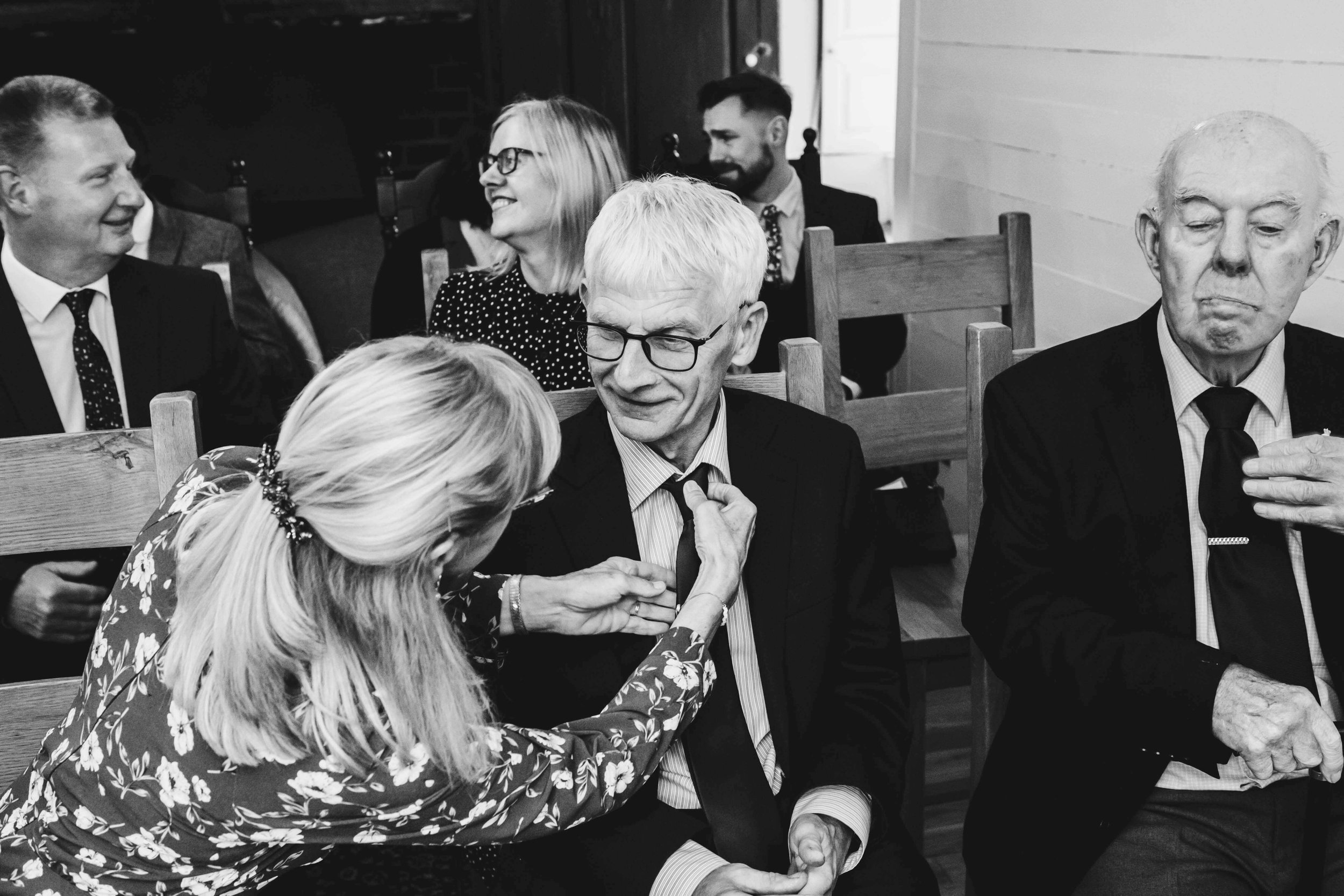 A woman with blond hair and floral dress pinning a badge on a blond man with glasses and a suit, while other people sit and talk in the background inside a wooden-paneled room.