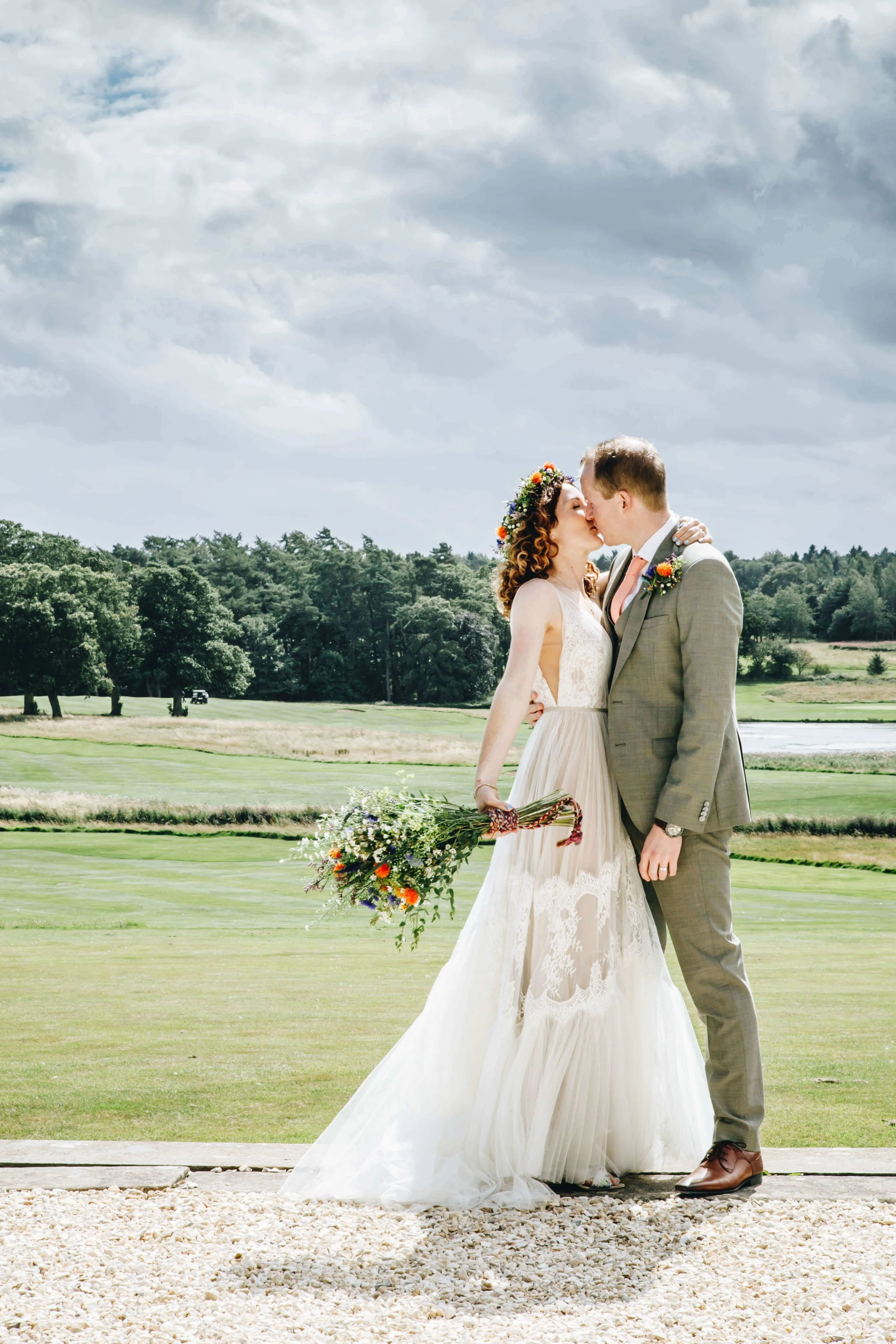 A bride and groom share a kiss outdoors on a wedding day, with a scenic countryside background and cloudy sky. Matfen Hall wedding photograph