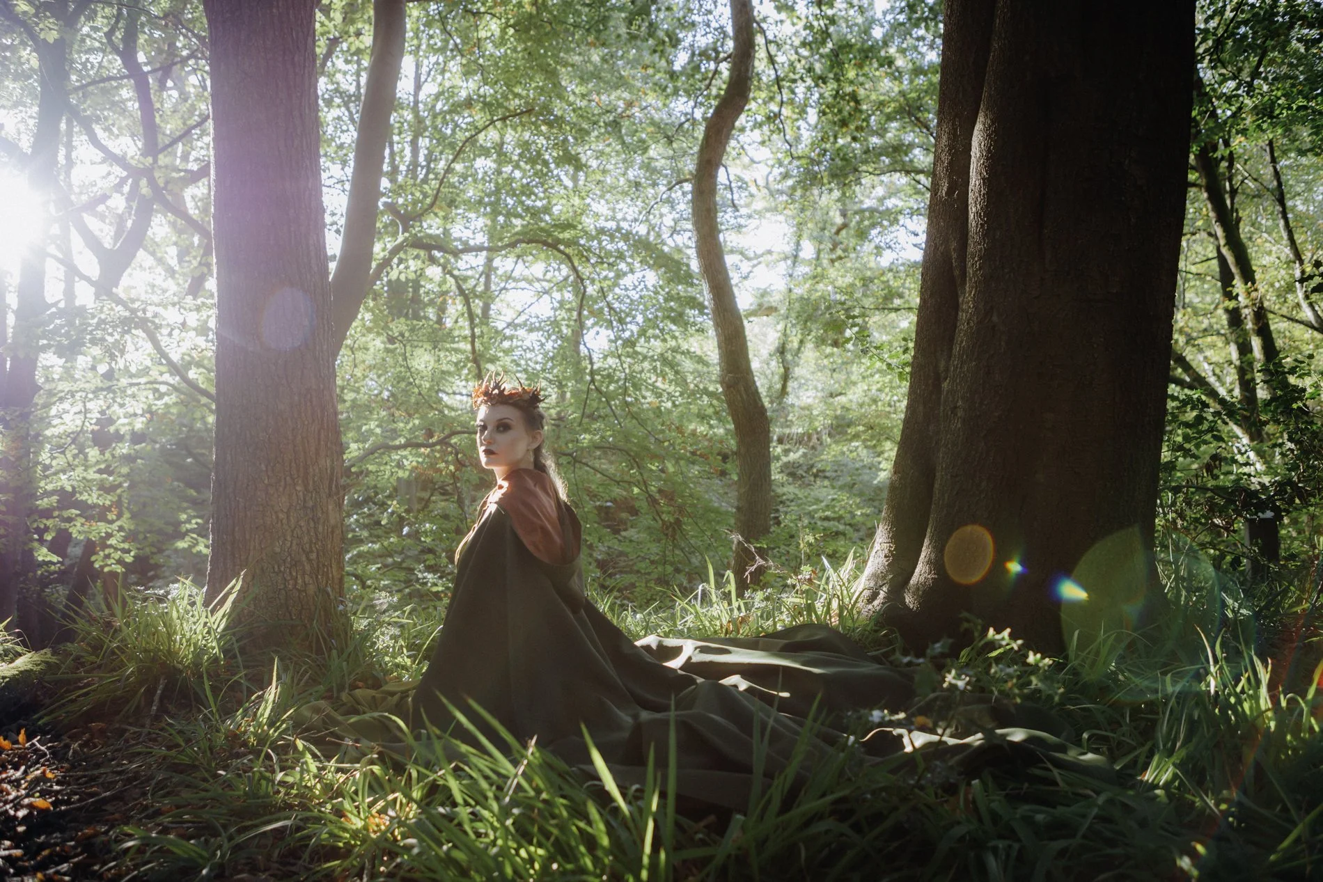 A woman sitting on the forest floor beside a large tree, wearing a dark cape with a hood, illuminated by sunlight filtering through the green leaves overhead.